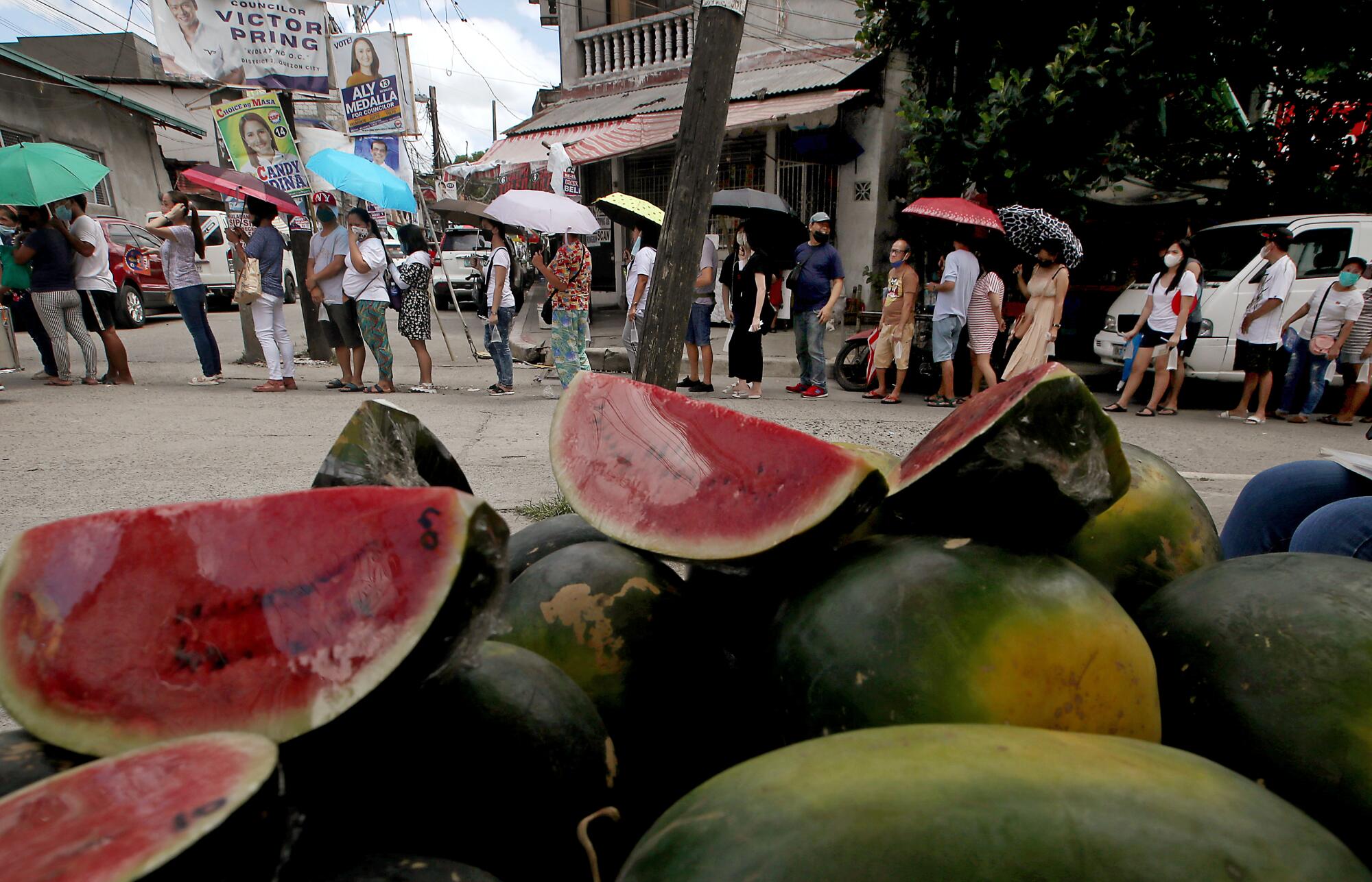 People lined up to vote, some holding umbrellas. In the foreground, watermelons for sale