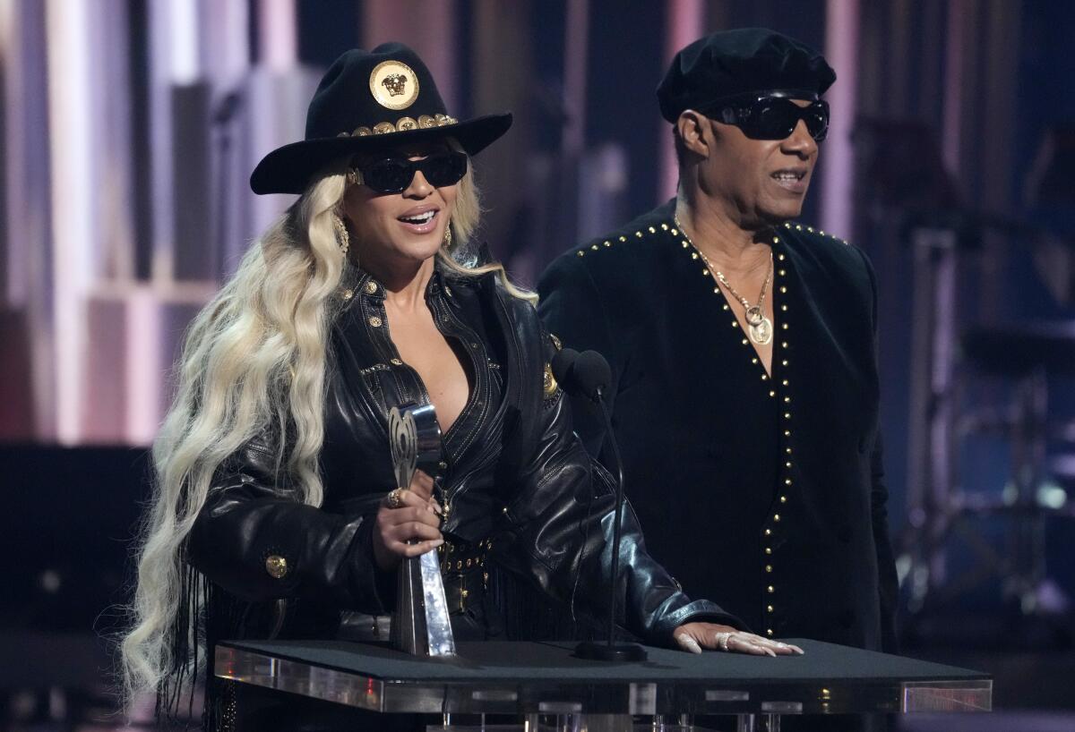 Beyoncé and Stevie Wonder both wear hats and dark glasses and stand together at a lectern as she holds an award