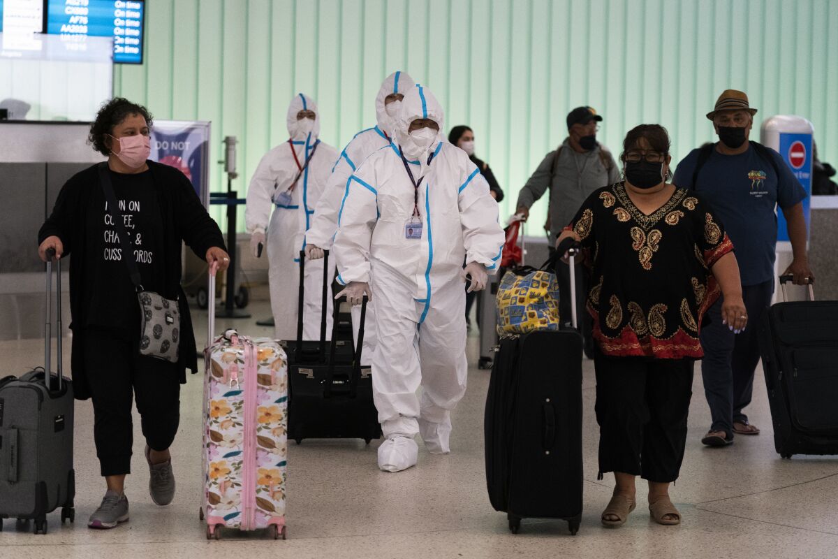 People in white coveralls, face coverings and plastic gloves pull their luggage alongside other fliers inside an airport.