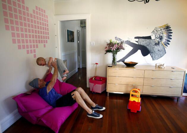 Scott Flora, on a bright pink Fatboy beanbag chair, lifts daughter Aja in the living room. The "Heart Breakout" decal above them was conceived by Hybrid Design and produced by Flora's company, Blik. "No one was really open to putting up something pink," Flora says. "I had no problem with it. I probably pulled and replaced almost all of the squares. We encourage people to play with the designs." To the right: David Bray's decal titled "I Never Saw the Sign" sits atop a Room & Board dresser used as sideboard near the dining area.