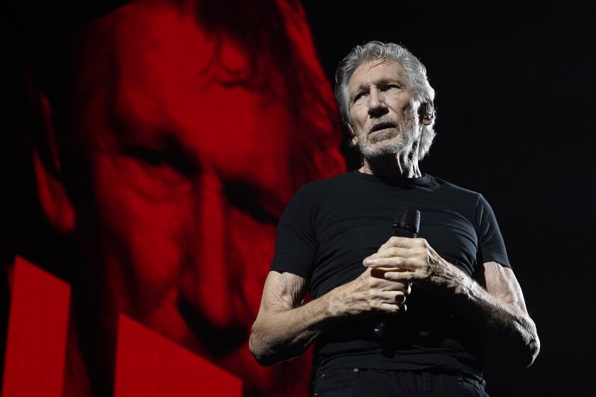 Roger Waters stands onstage holding a microphone with two hands as a face is projected in red behind him