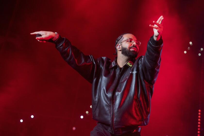 Drake in a large black leather jacket and glasses holding his hands out in front of him on a stage
