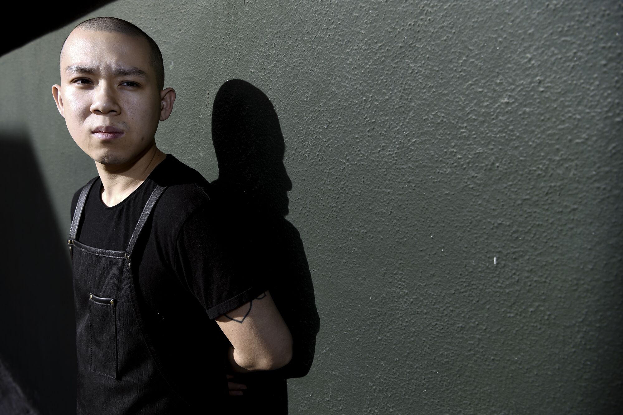 A person dressed in black shirt and chef's apron leans against a black wall.