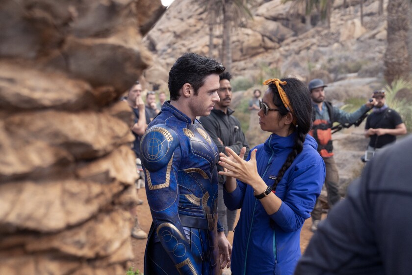 A woman in a blue jacket and sunglasses, right, talks to a man in a blue superhero suit.