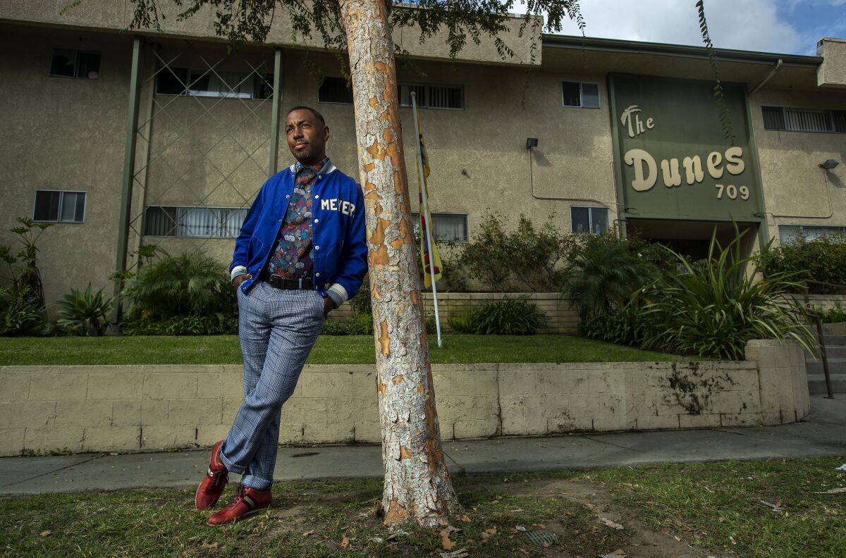 A man in blue jeans and an athletic jacket leans against a tree outside an apartment complex