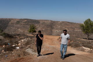 A man in dark clothes, left, and another man, in a white shirt and jeans, walk on a dirt path