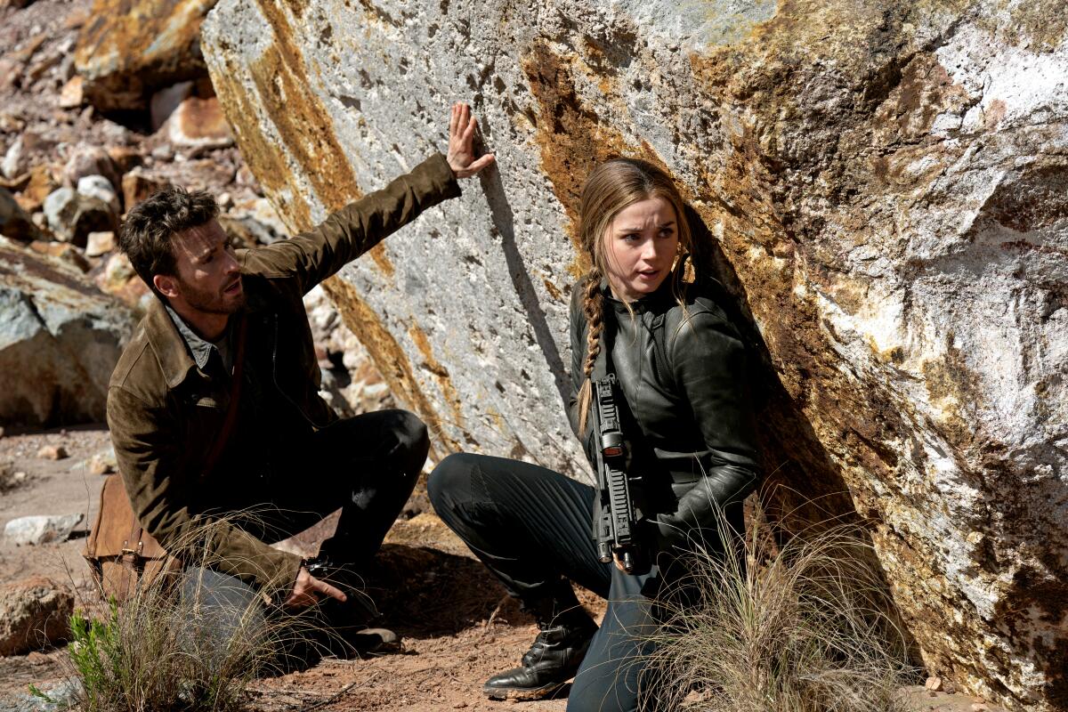 A man and a woman with a gun crouching beside a rock.