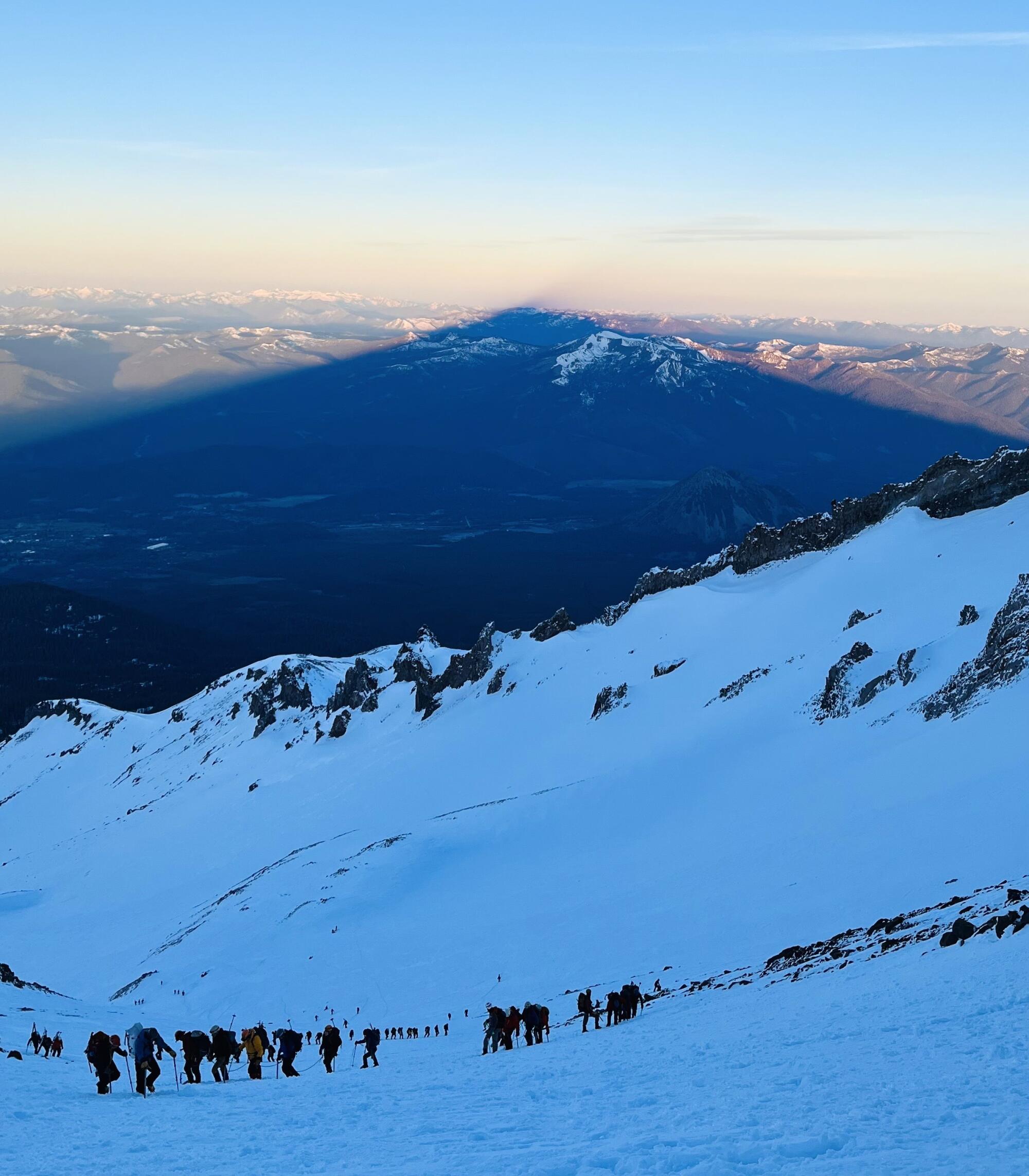 Groups of climbers on a snowy mountainside as a triangular shadow stretches into the distance.