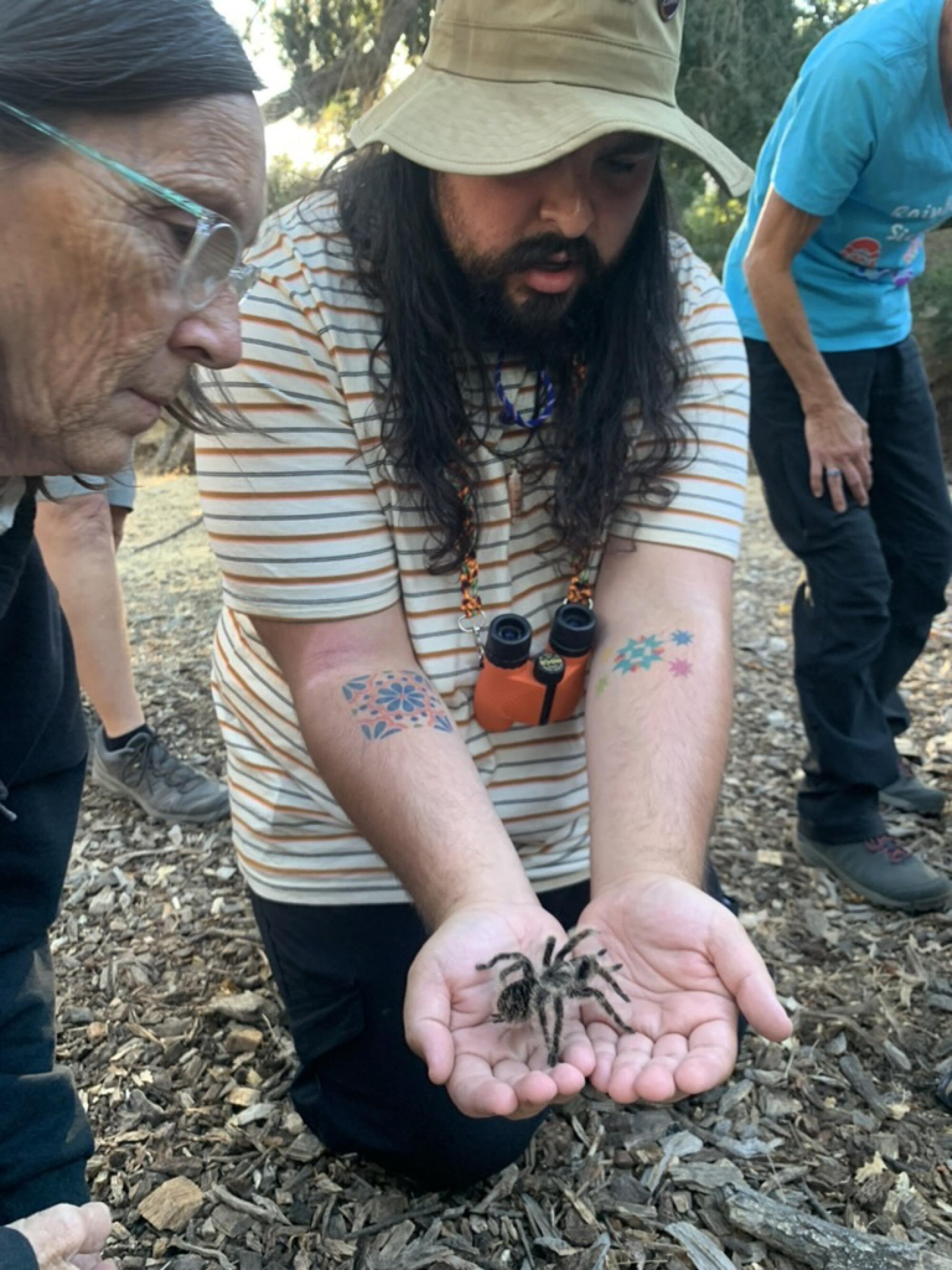 A man kneeling on the ground, holding out a tarantula in his cupped hands as others look on