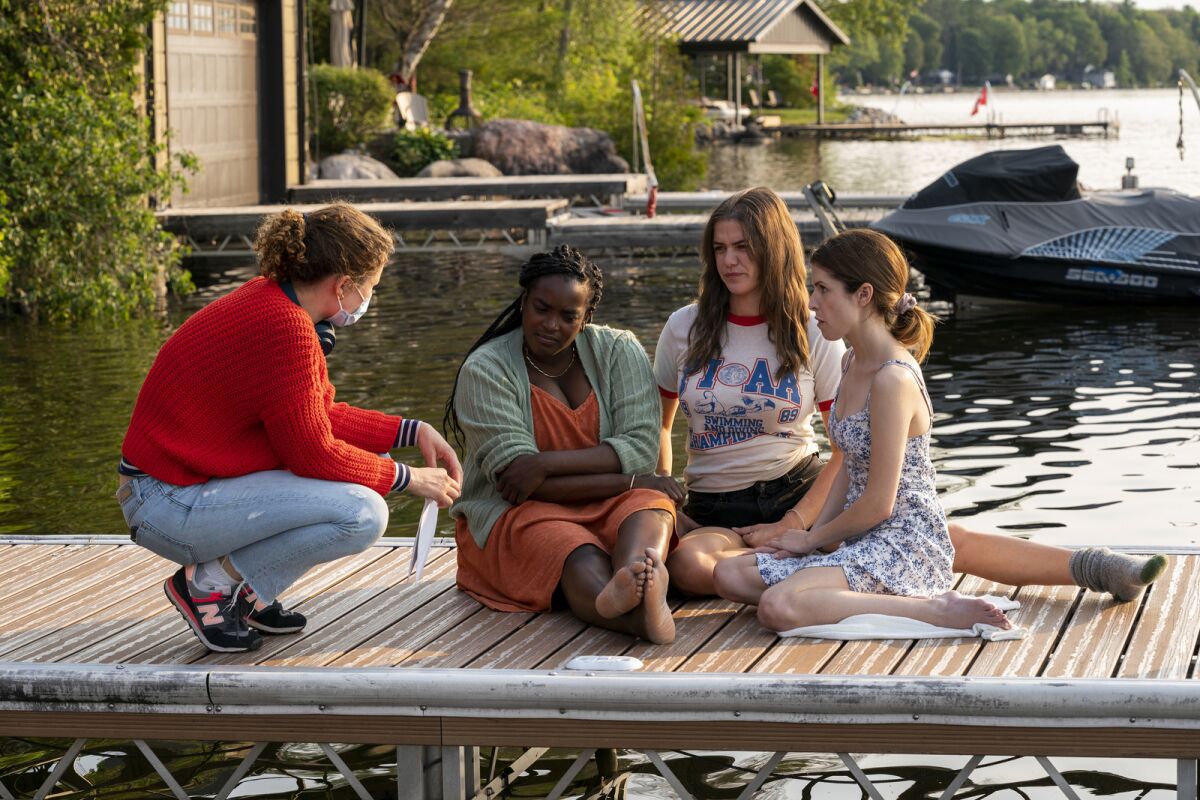 A woman director kneels to talk with three actors who sit on a dock.