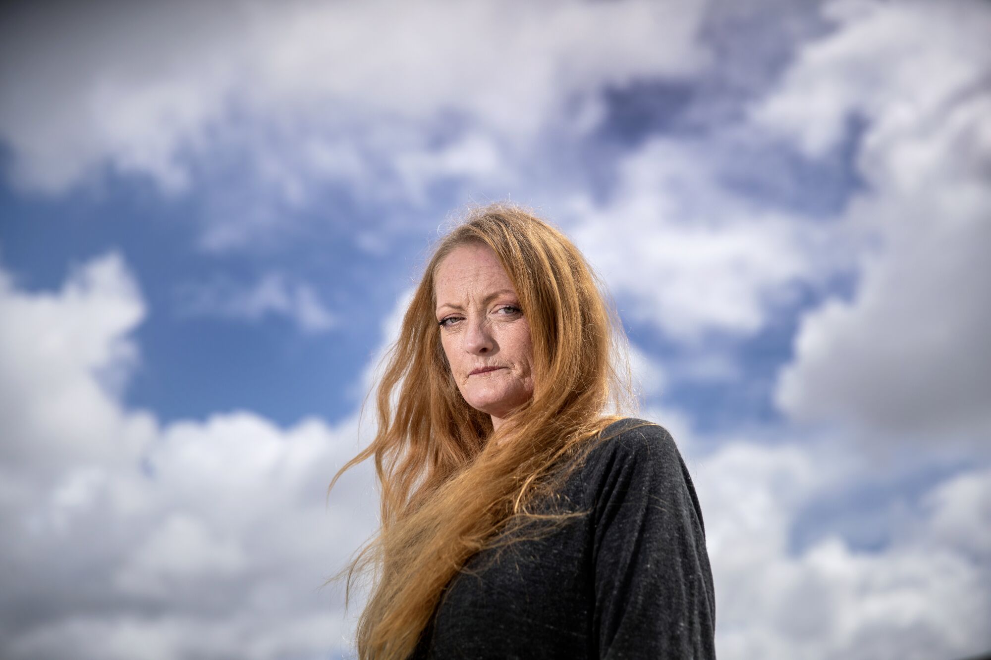 A woman with long copper-toned hair looks at the camera. Sky with fluffy white clouds fills the background.