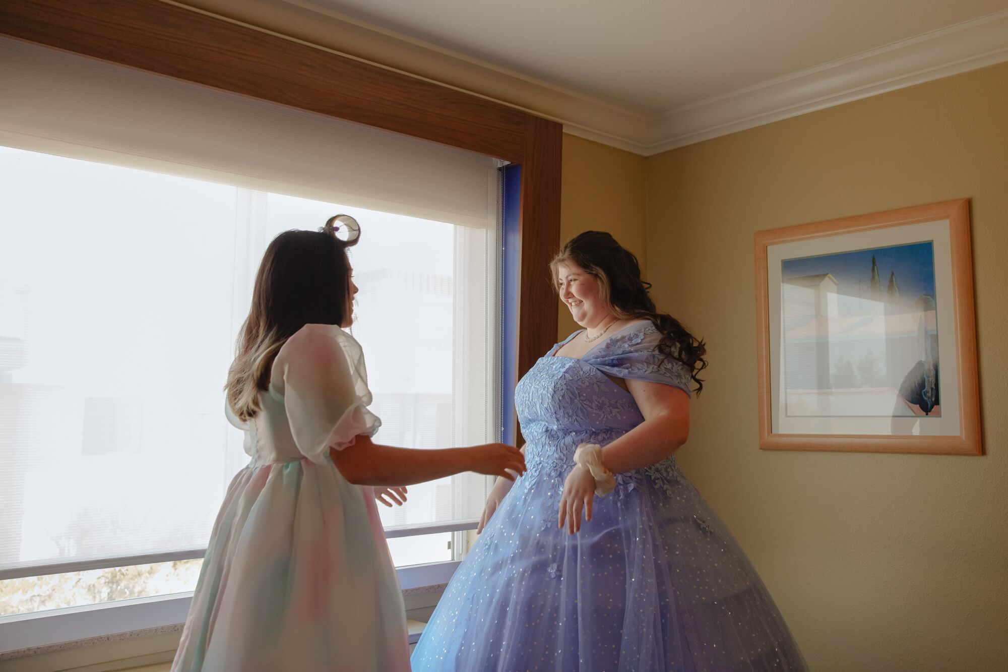 Two women in ball gowns stand in front of a window.