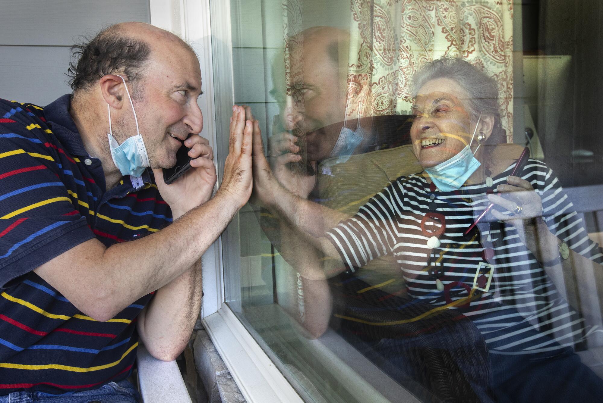 A man and his mother raise their palms to each other from opposite sides of a window while talking on cellphones