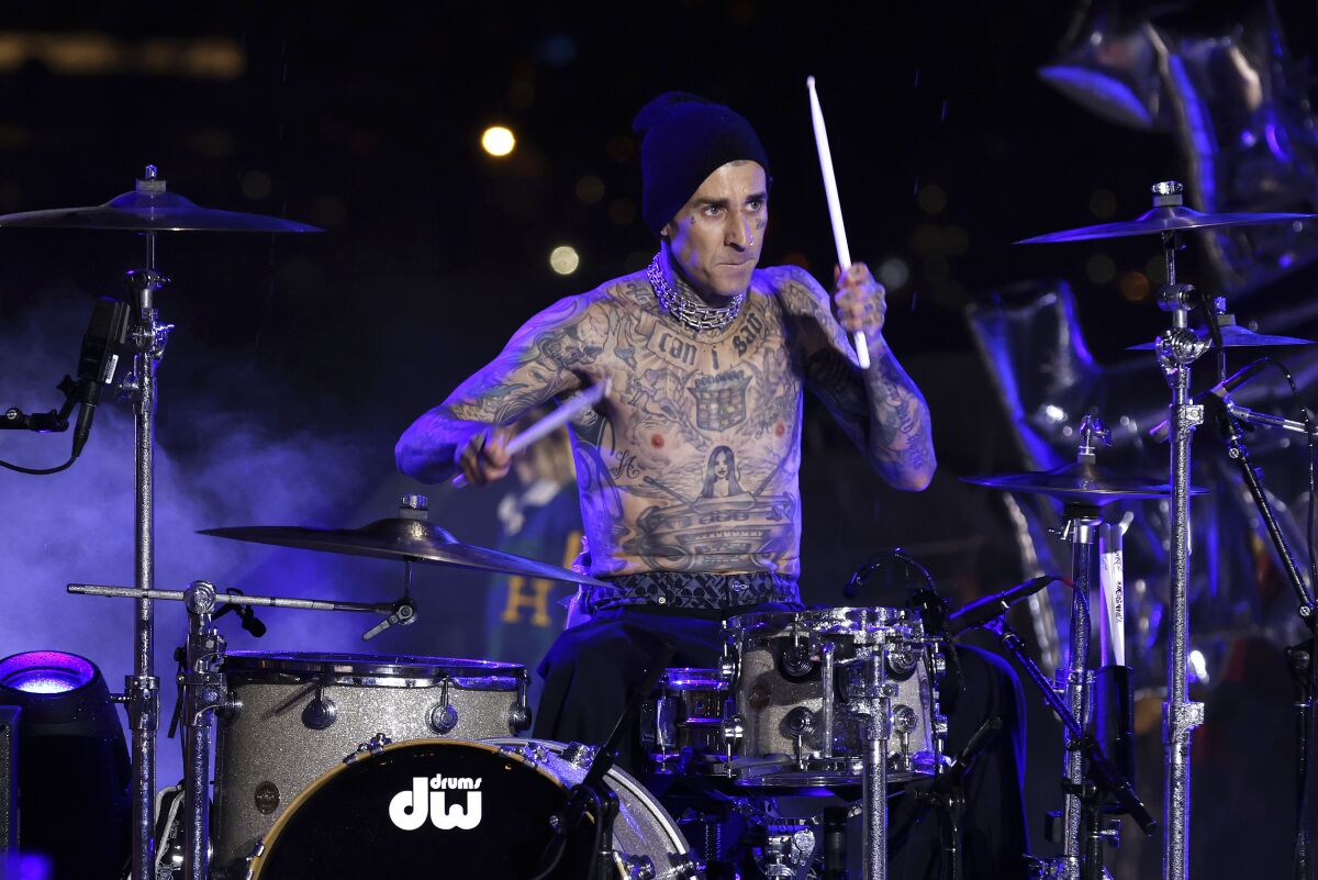 A shirtless man with tattoos all over his body wearing a black beanie and playing the drums