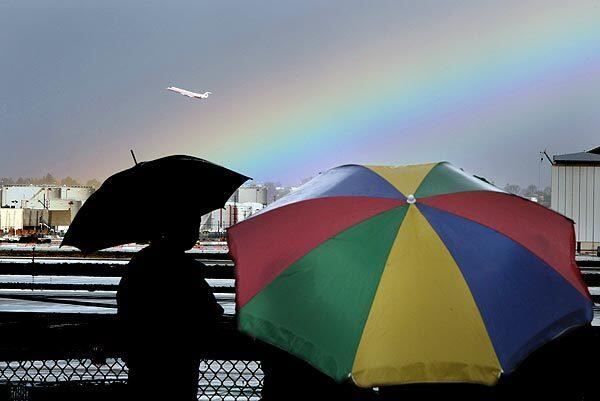 Families huddle beneath umbrellas to watch a rainbow as a jet takes off from Los Angeles International Airport. After a week of severe storms in the Southland, milder weather is expected.