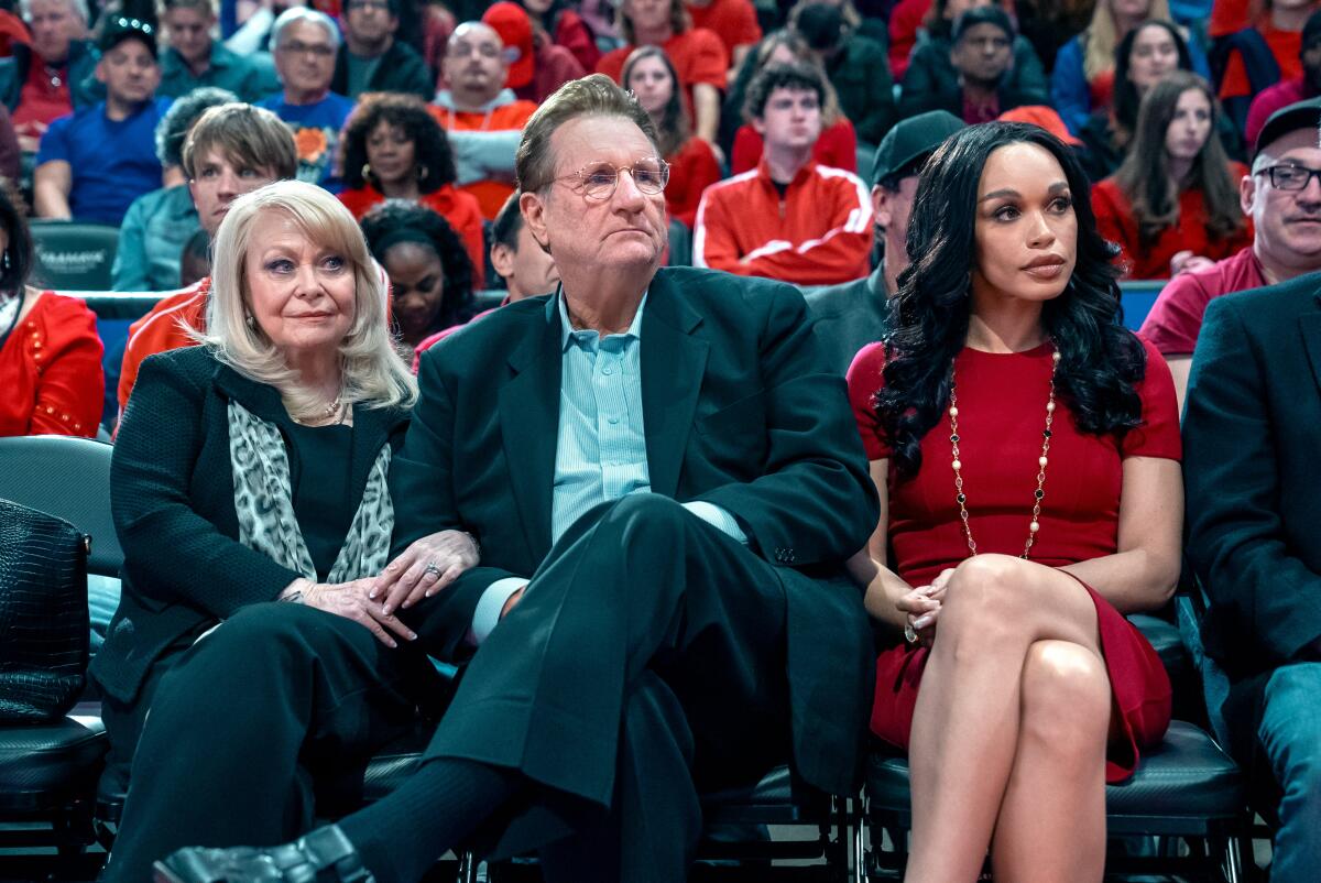 A man sits courtside at a basketball game with two women flanking him.