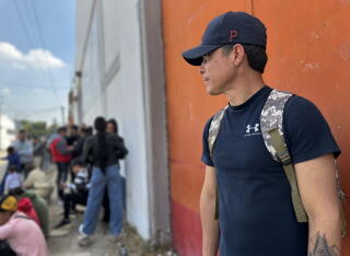 A man in a dark T-shirt and cap stands in front of an orange-colored wall, looking at people milling about nearby