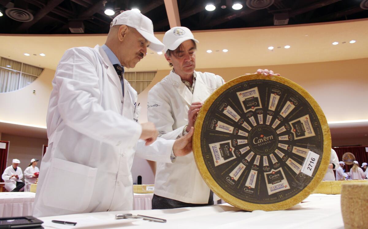Two men in white inspect a wheel of cheese.