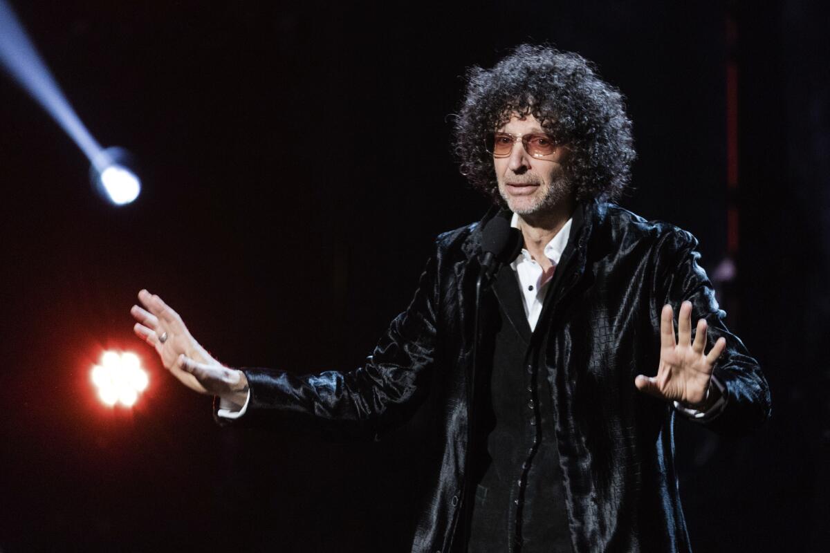 A man with voluminous, curly hair in a shiny black suit speaks onstage