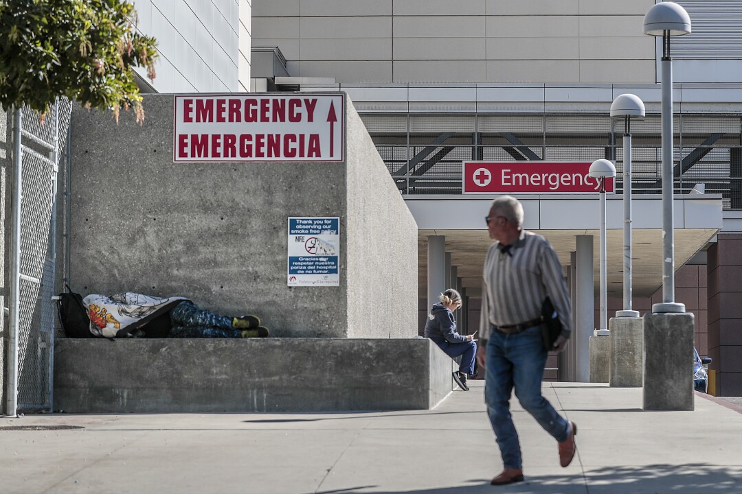 People sit and lie down on a concrete bench outside a hospital as another man walks by