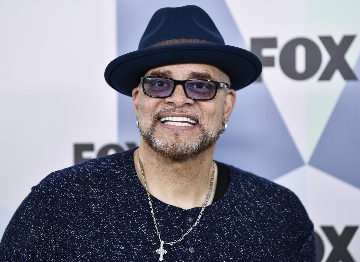 Sinbad in a black wide-brimmed hat, black shirt, necklace with a cross pendant and sunglasses smiling