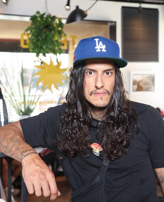 Actor Richard Cabral poses at a coffee shop. He wears a black shirt, has long black hair, and has a Dodgers hat on. 