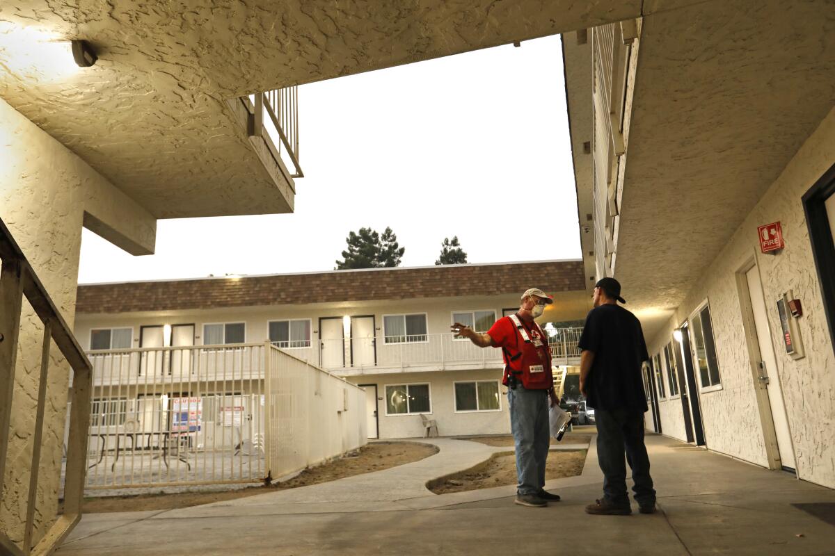 A Red Cross volunteer in a mask and reflective vest talks to a fire evacuee in the courtyard of a motel