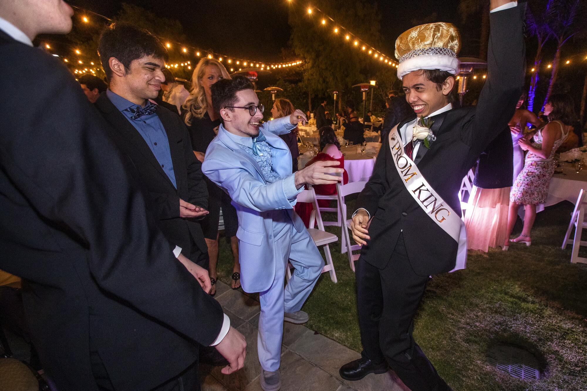 Teenagers in suits dance while one wears a crown and "Prom King" sash