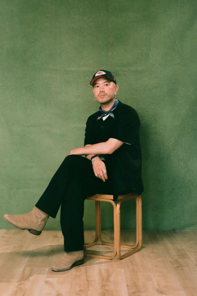 Caleb sits with his legs crossed on a stool in front of a green backdrop