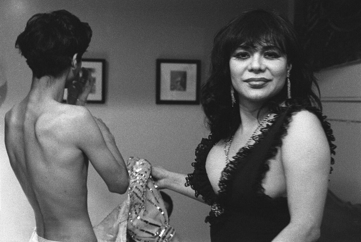 A trans woman in a ruffled dress smiles at the camera as a shirtless figure is seen behind her.
