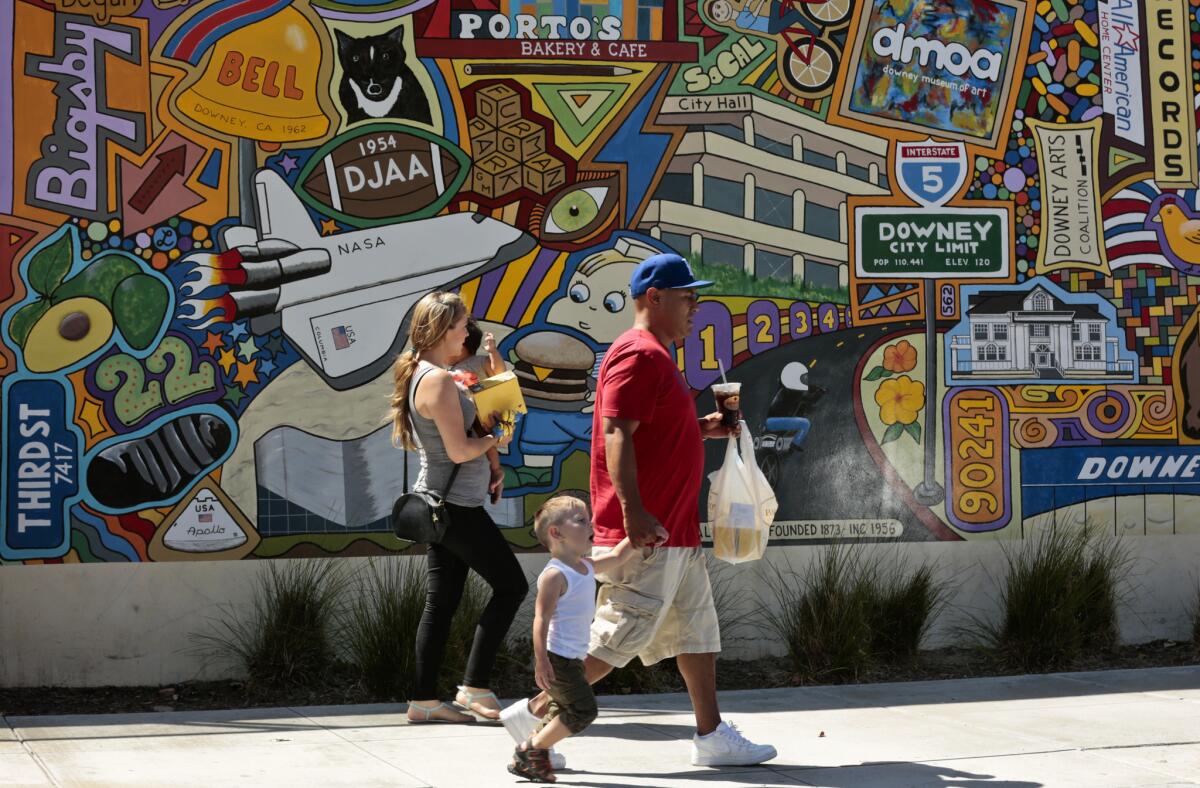 A family walks past a mural on Downey Avenue with their food from Porto's Bakery and Cafe. The mural depicts the diverse middle-class suburb of Downey, former home of the Space Shuttle.