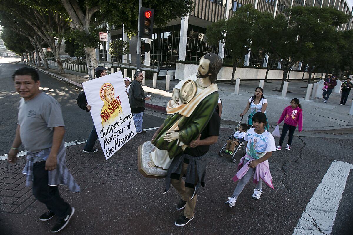 A man carrying a statue passes members of the Mexica Movement, on their way to protest the sainthood of Father Junipero Serra at the gates of the Cathedral of Our Lady of the Angels in downtown L.A.