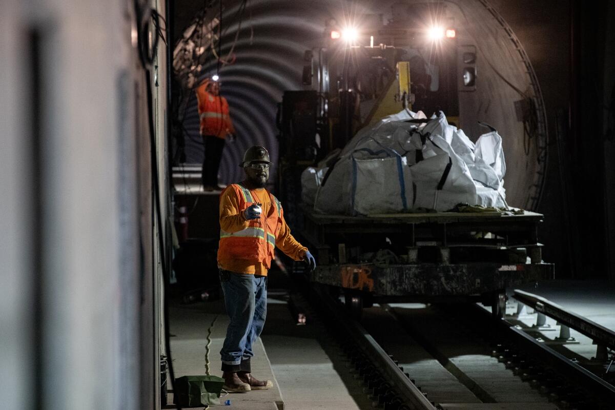 Two workers in orange safety gear alongside a train car carrying materials in a tunnel