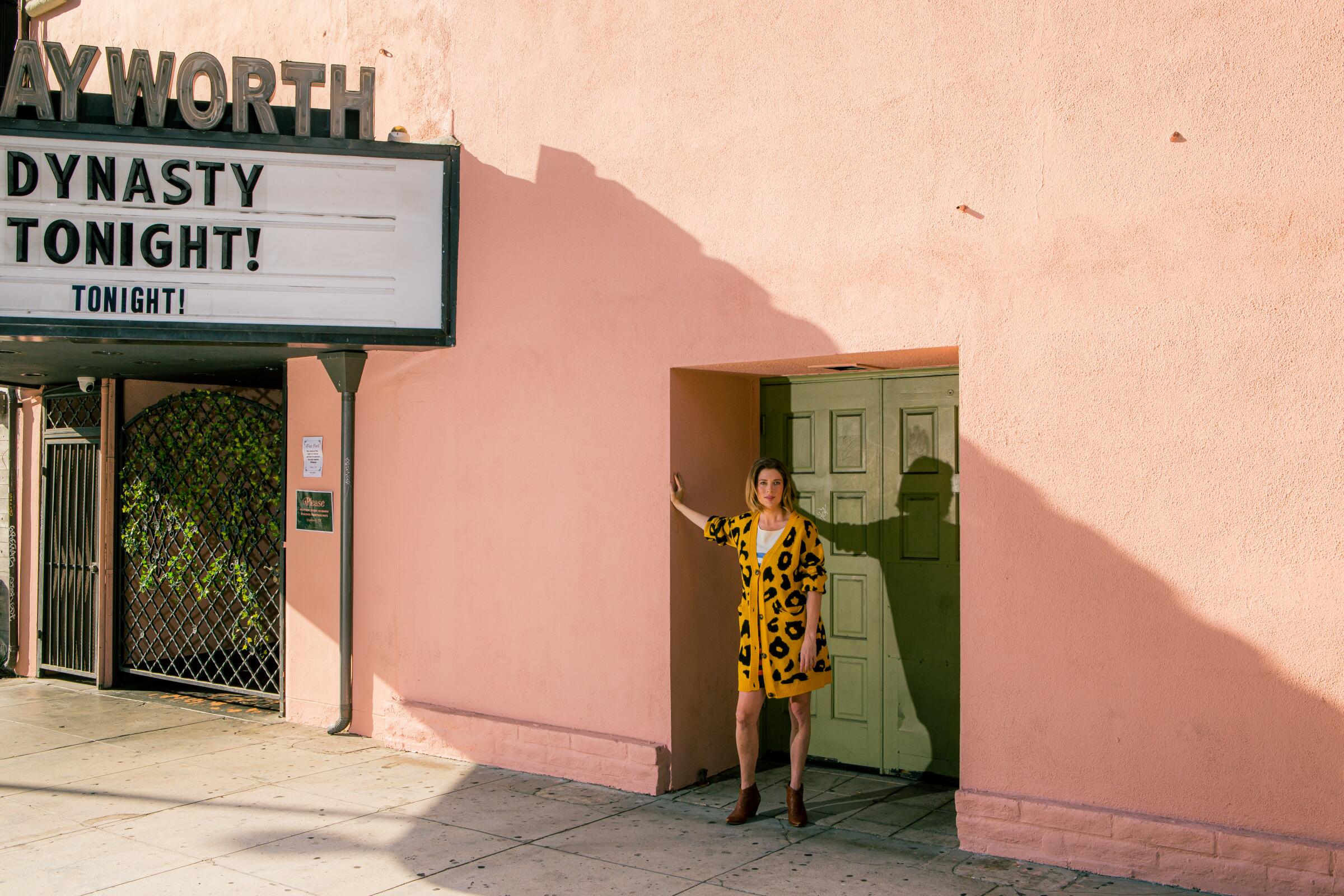 A woman stands outside the doorway of a building, with one arm against a wall