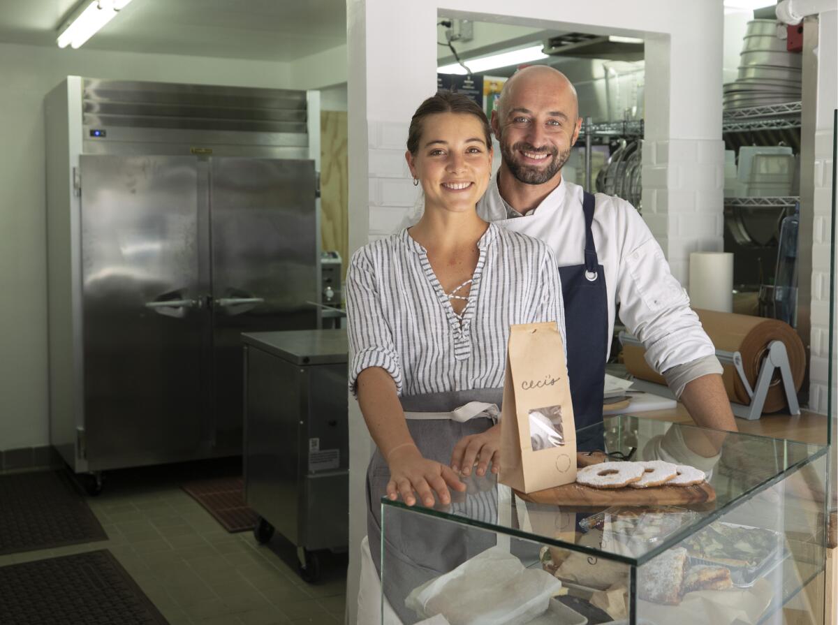 A man and a woman in aprons smile in a kitchen 