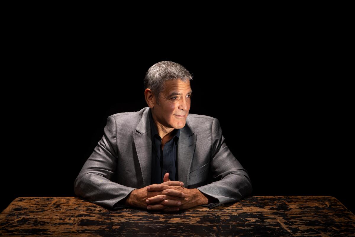 A light cuts through darkness, showing George Clooney sitting, resting his hands, fingers laced, on a distressed wood surface