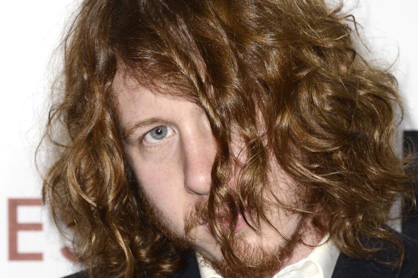 A man in a suit looks coyly out from behind his wavy red hair as it falls over his face