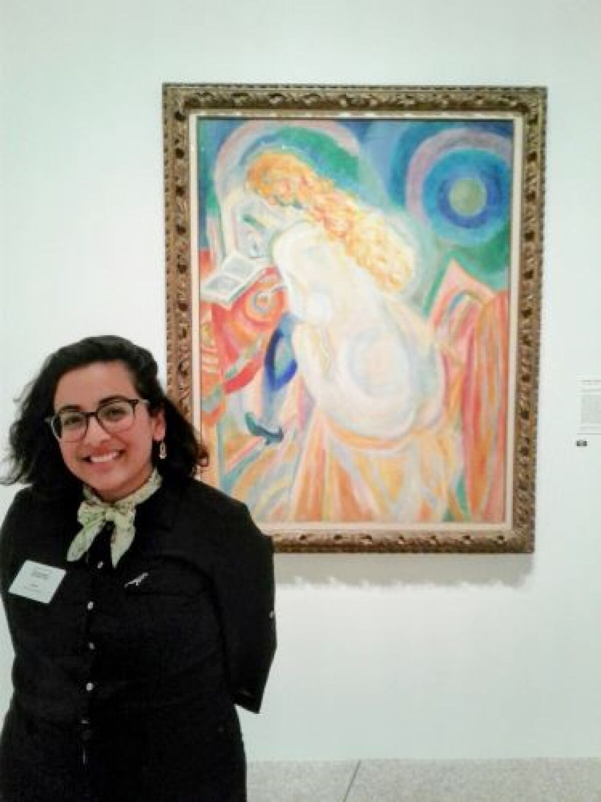 A woman in a uniform and name tag stands, smiling, in front of a painting in a museum