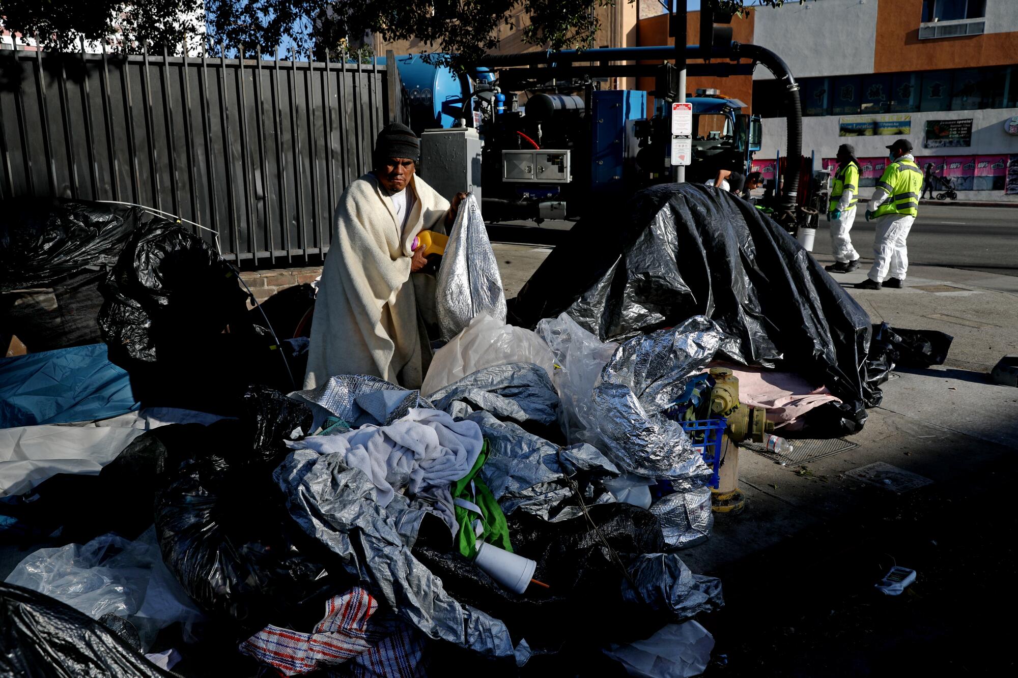A man wrapped in a blanket stands on a sidewalk surrounded by bags.