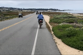 A gif of a woman bike riding along a beach road.