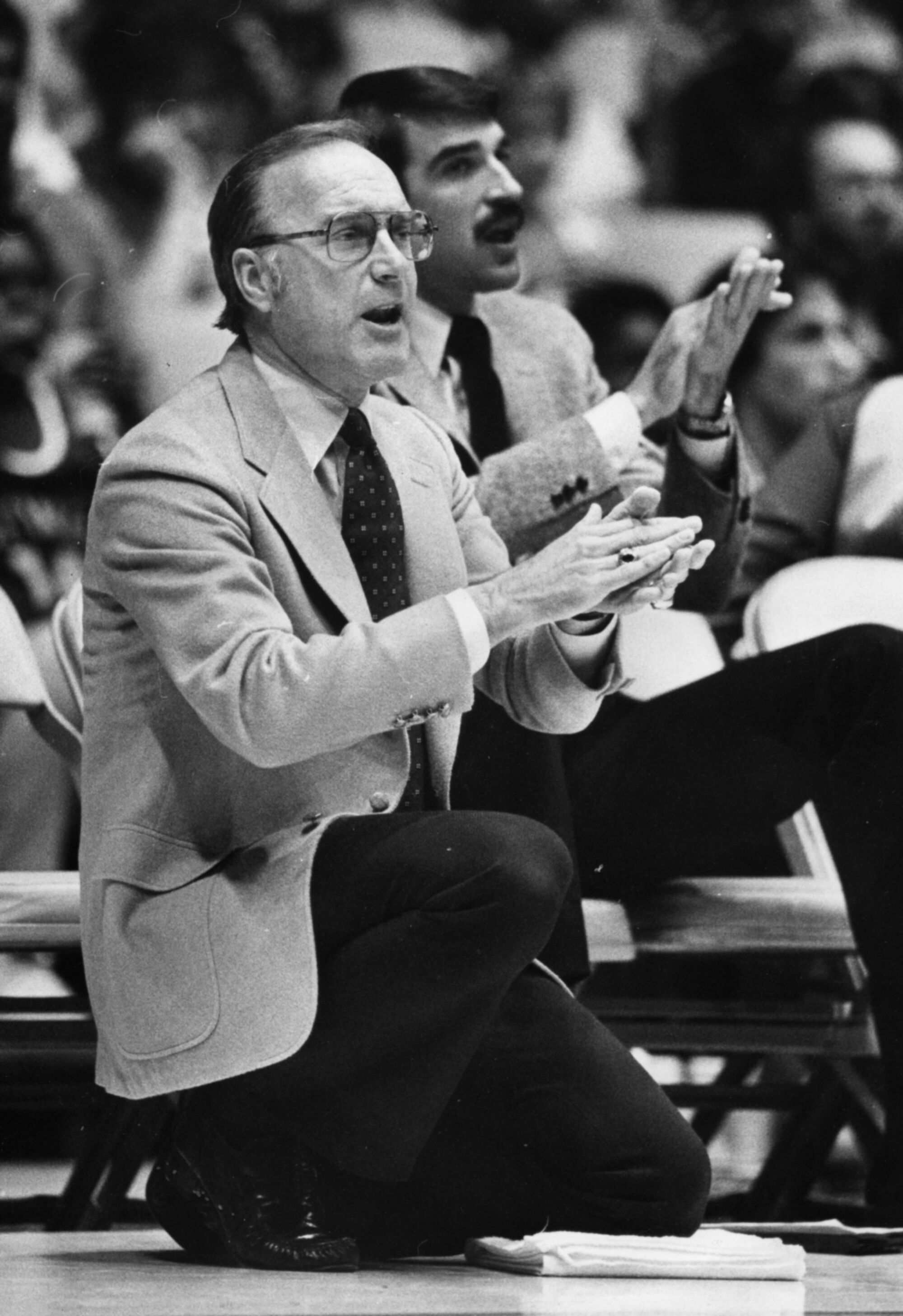 A man in glasses claps while kneeling on the basketball court sideline