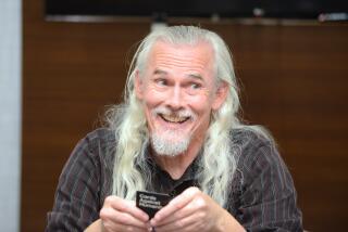 A man with long silver hair in a dark long-sleeve shirt holding a black card in his hands and smiling 