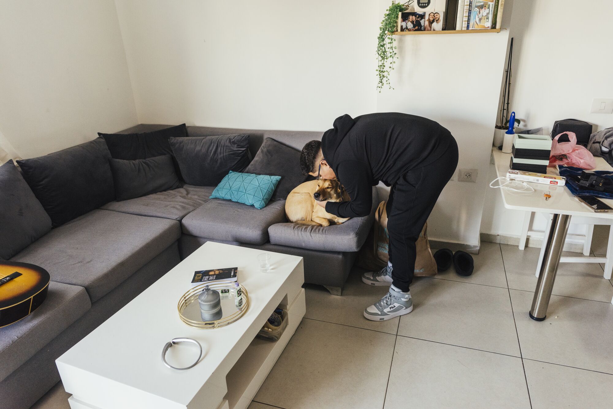 A man with dark hair, wearing dark clothes, bends down to kiss a dog on a couch 