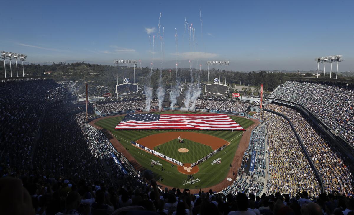 A distant aerial view of a packed baseball stadium with a huge U.S. flag stretched across the outfield.