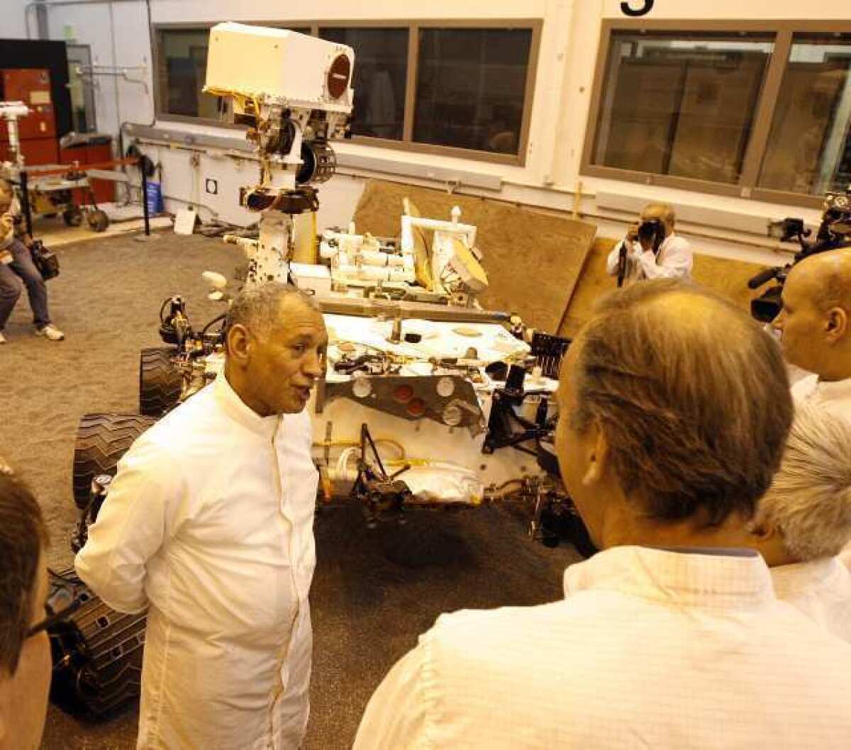 NASA Administrator Charles Bolden talks with scientist and engineers at JPL next to a duplicate of the Mars Rover Curiosity at JPL. The room is bathed in light that is similar to the light on the martian planet.