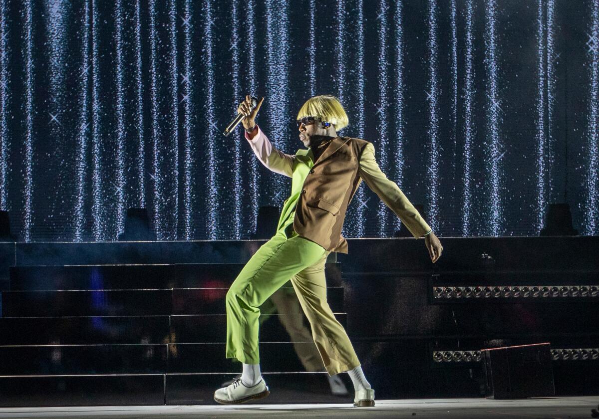 Tyler, the Creator in a platinum blond bowl cut wig and a green and brown suit posing on a stage