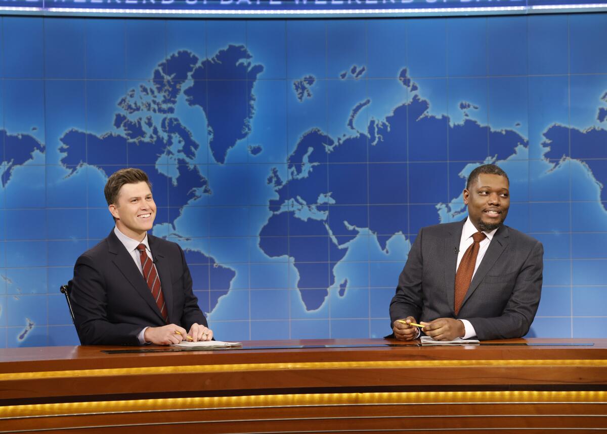 Colin Jost and Michael Che wearing suits and sitting behind the "Weekend Update" desk on "Saturday Night Live"
