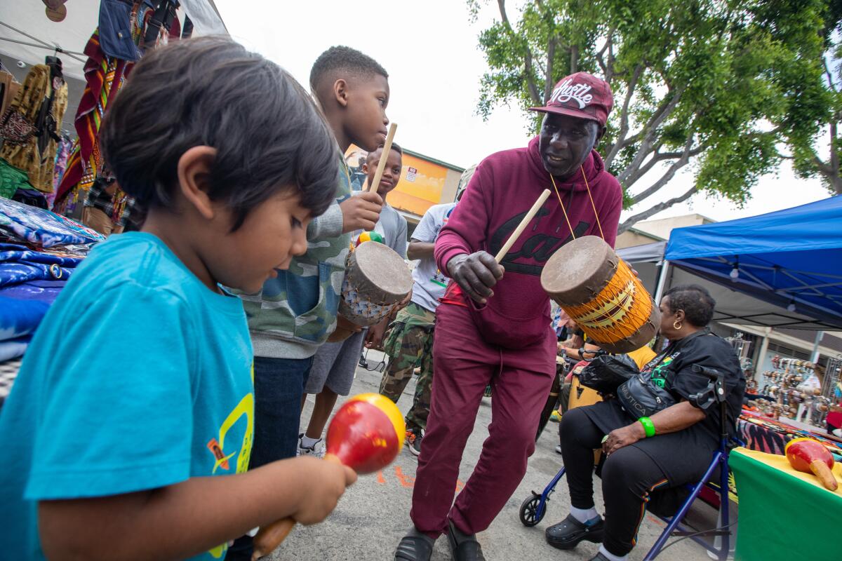 A man beats a handheld drum while children hold a drum and a shaker while outdoors at a fair. 