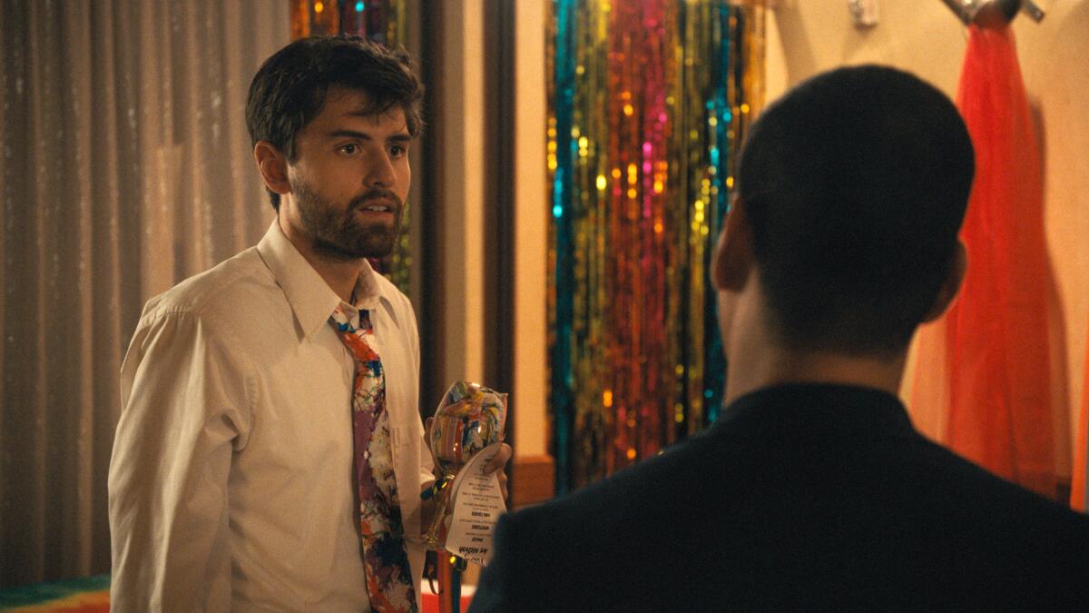 A man in a collared shirt and bright tie stands in a banquet room talking to another man. 