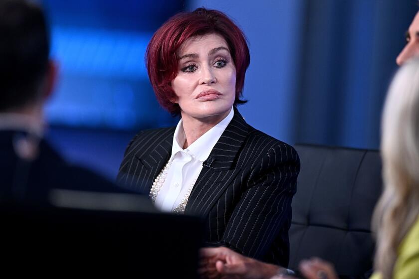 Sharon Osbourne is sitting on a panel, listening with a serious face, wearing a black pinstripe suit and white shirt